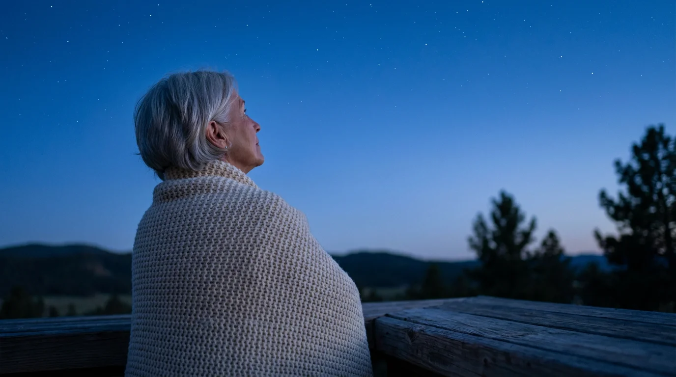 A senior woman wrapped in a blanket, stargazing from a deck at dusk.