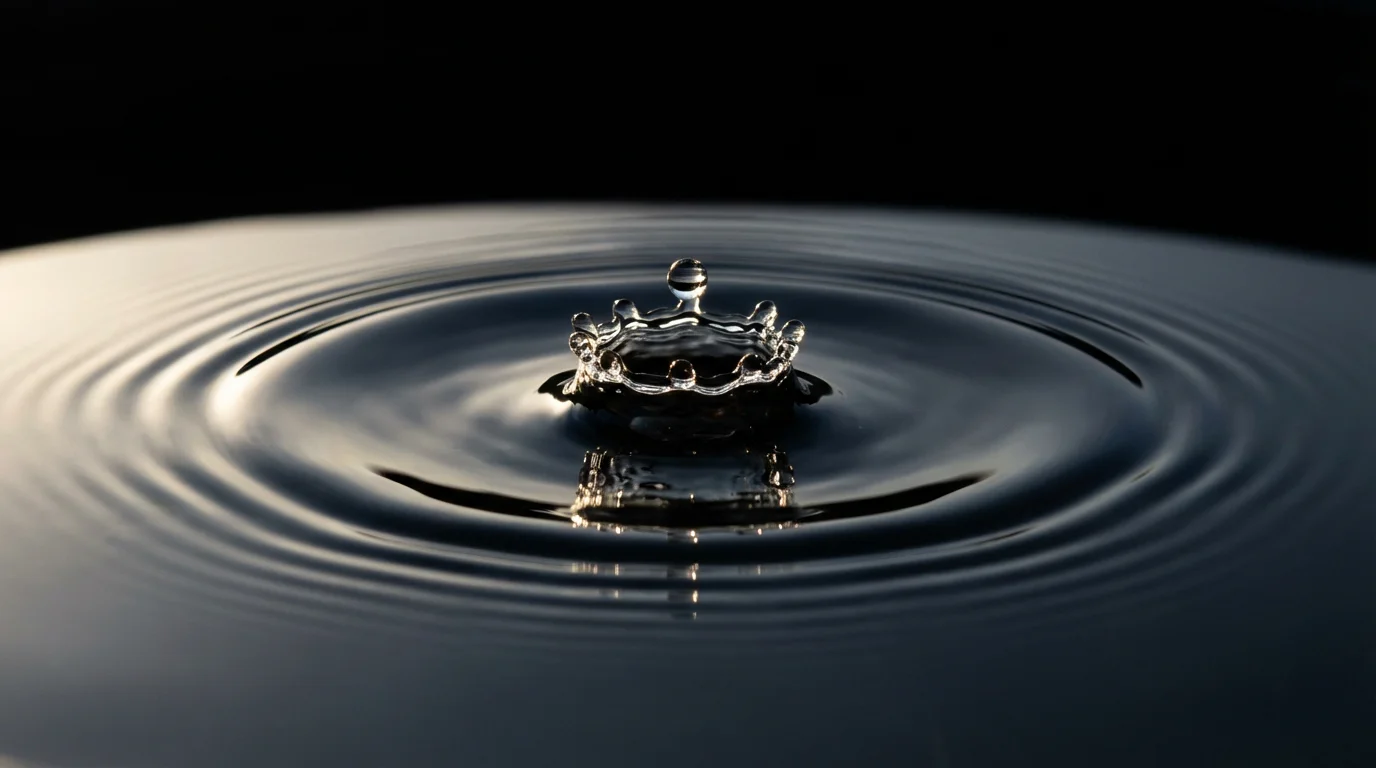 Macro photograph of a water droplet creating expanding ripples on a dark water surface.